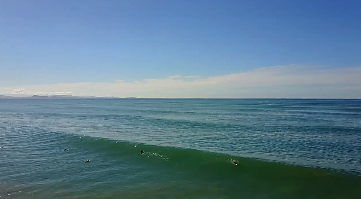 A few surfers stroke into glassy peaks under a big blue sky, Aramoana surf spot, Dunedin / Otago, New Zealand.